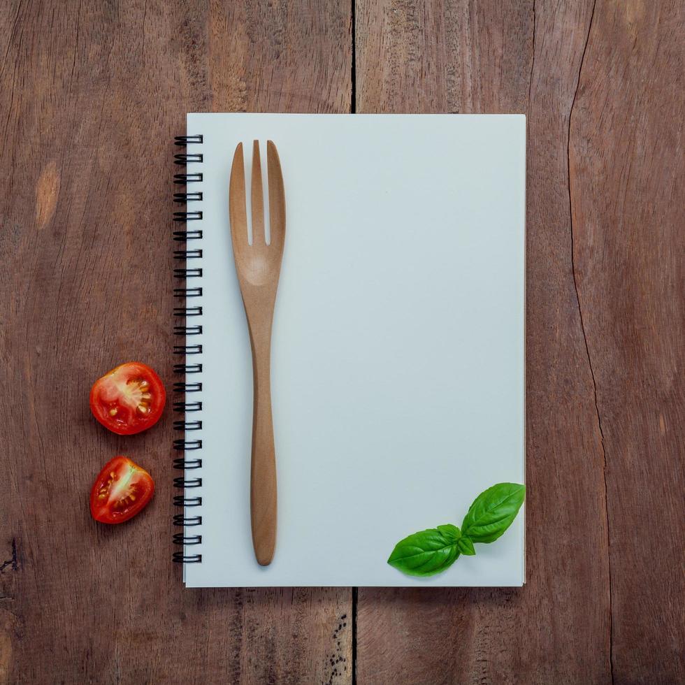 Cuaderno, albahaca dulce, rodaja de tomates cherry y tenedor sobre fondo de madera en mal estado oscuro foto