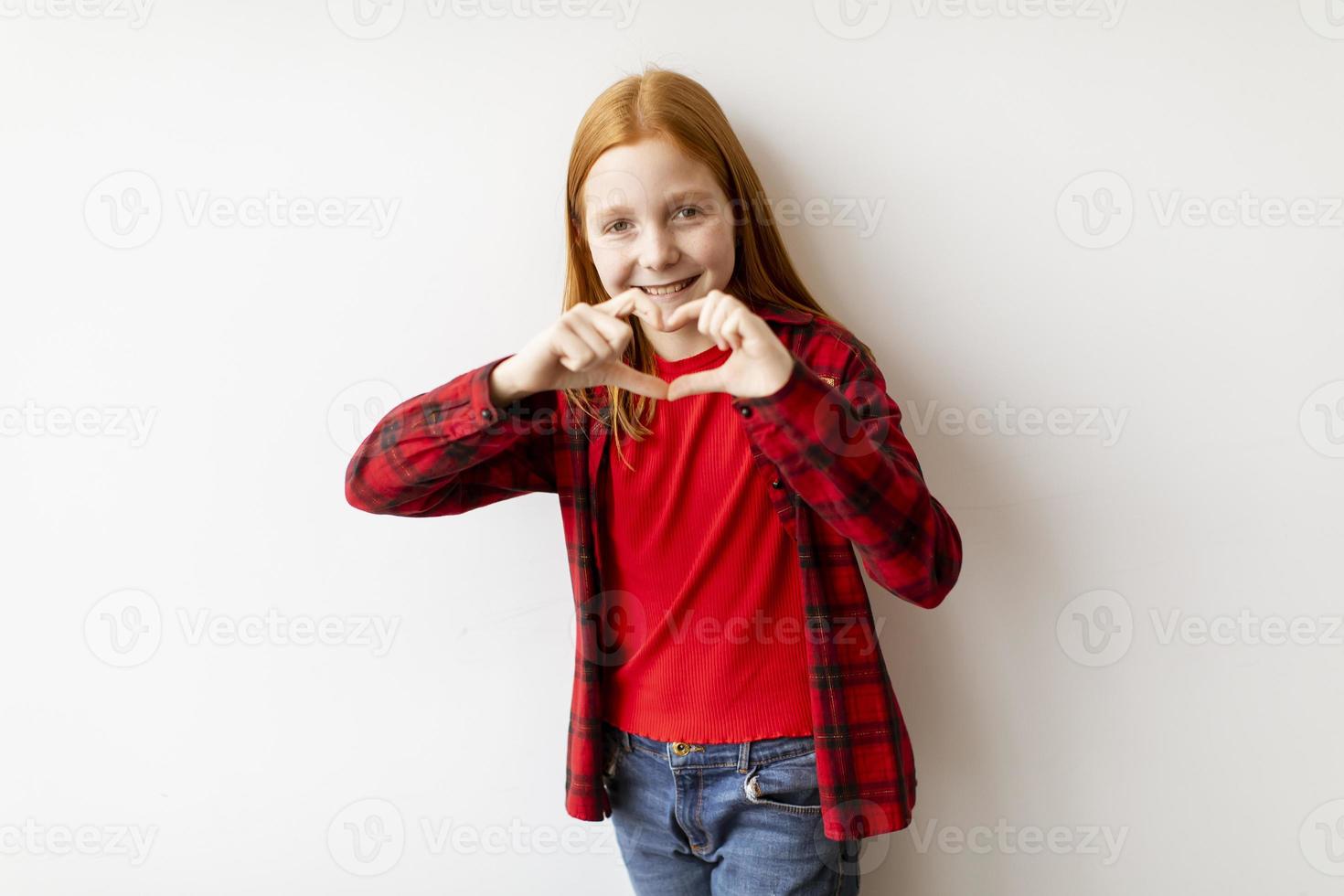 linda niña de pelo rojo de pie junto a la pared blanca y mostrando la forma de corazón con los dedos foto