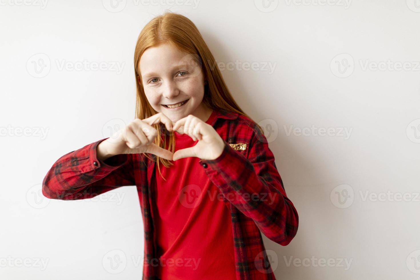 linda niña de pelo rojo de pie junto a la pared blanca y mostrando la forma de corazón con los dedos foto