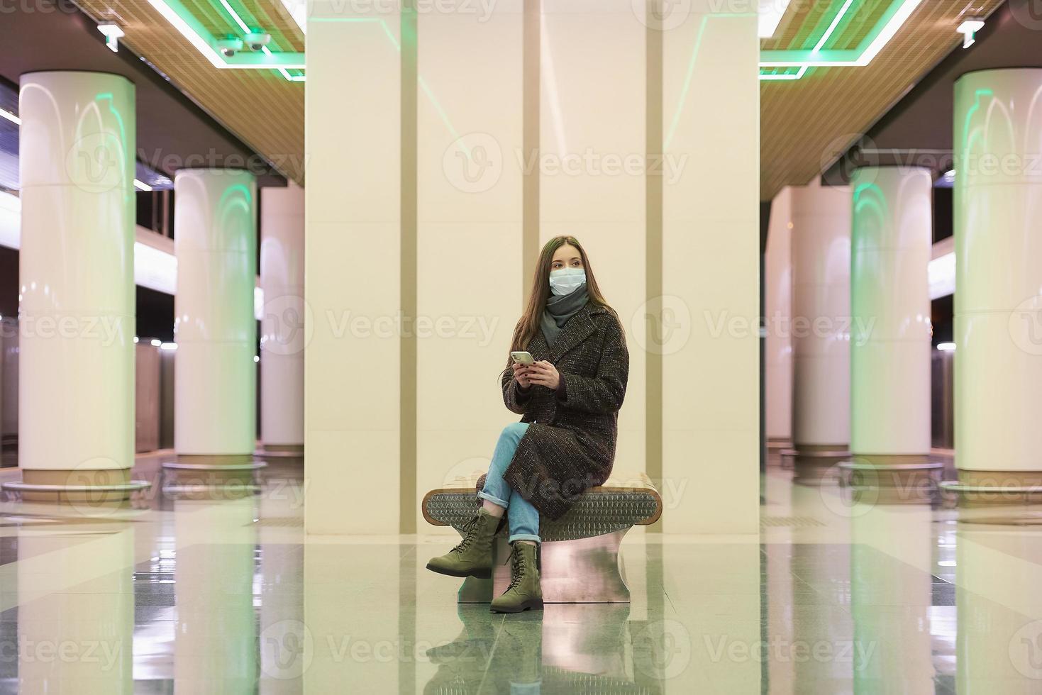 A woman in a medical face mask is waiting for a train and holding a smartphone photo