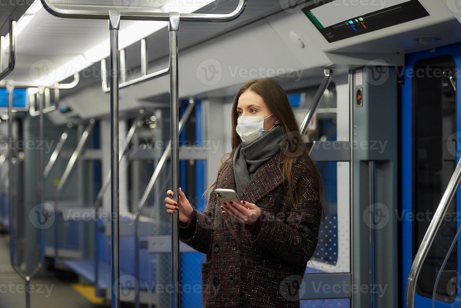 una mujer con una mascarilla está de pie y usando un teléfono inteligente en un vagón de metro moderno foto