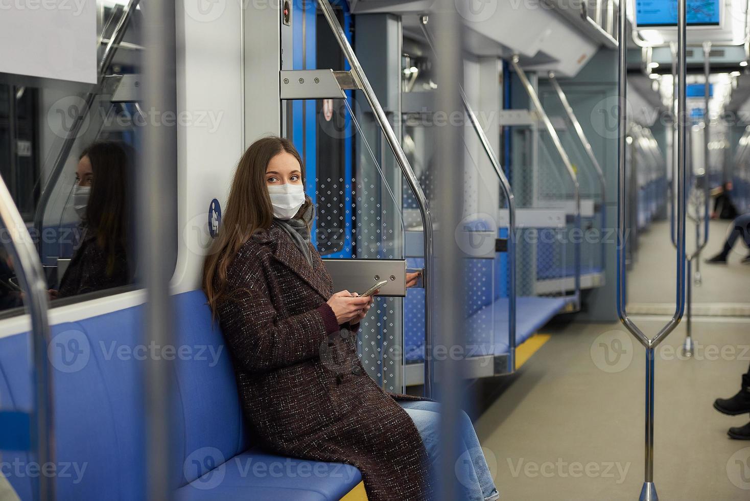 una mujer con una mascarilla está sentada y usando un teléfono inteligente en un vagón de metro moderno foto