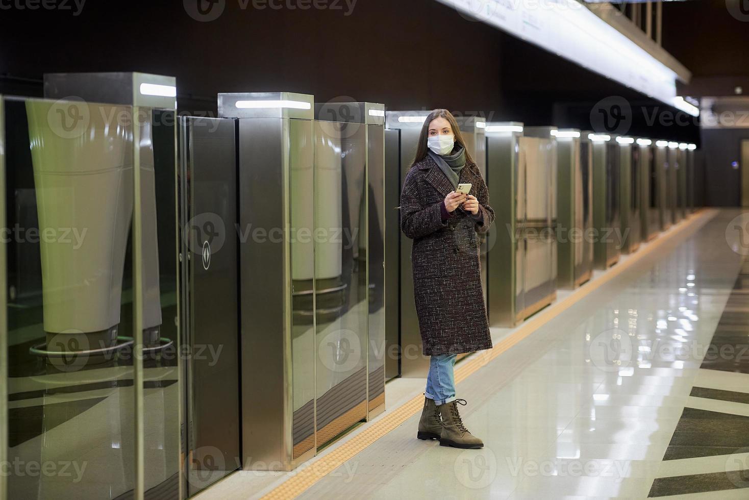 una mujer con una mascarilla médica está esperando un tren y sosteniendo un teléfono inteligente foto