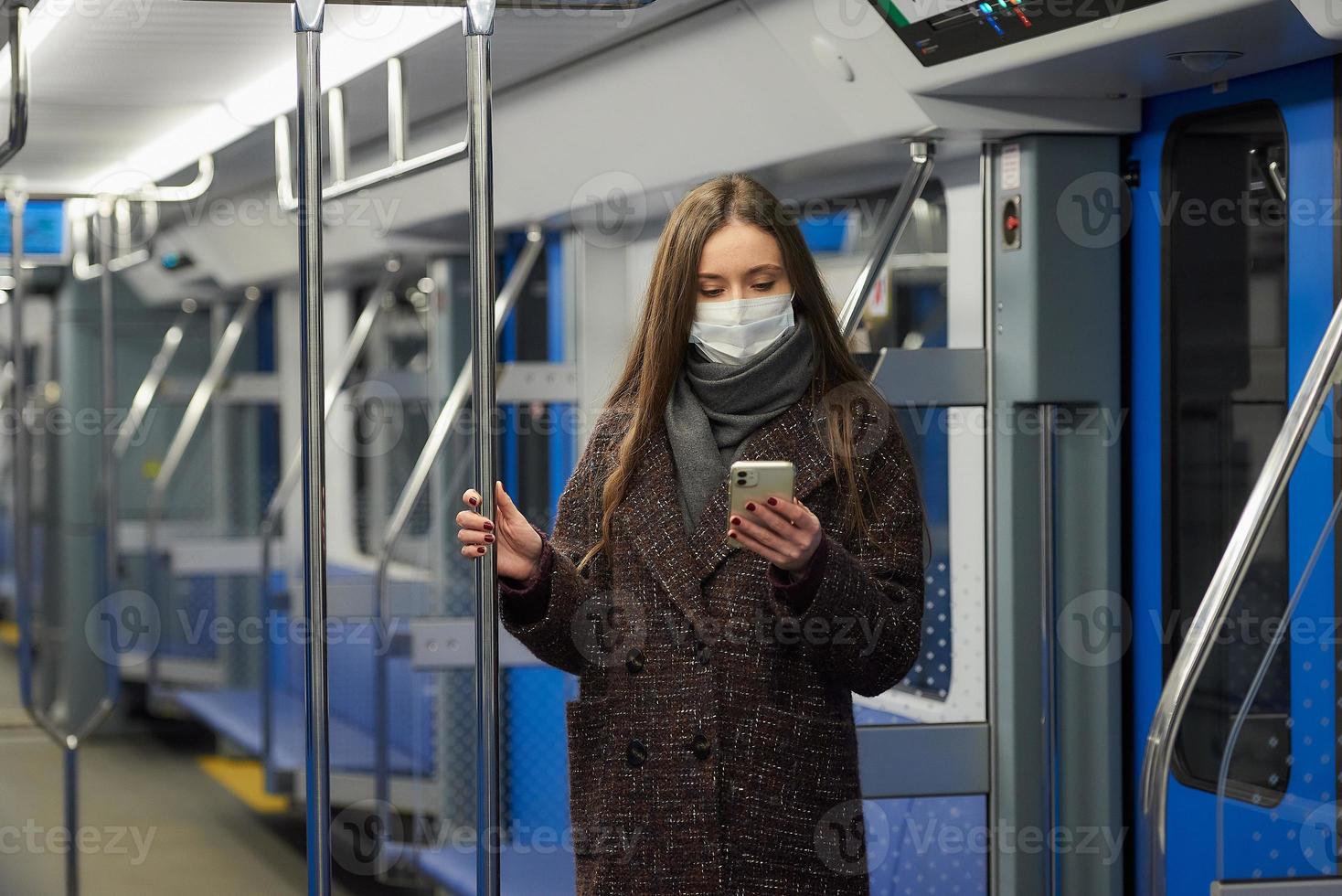 una mujer con una mascarilla está de pie y usando un teléfono inteligente en un vagón de metro moderno foto