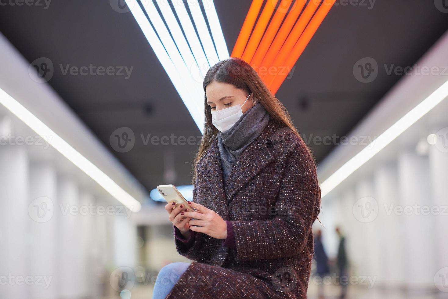 una mujer con una mascarilla médica está esperando un tren y sosteniendo un teléfono inteligente foto