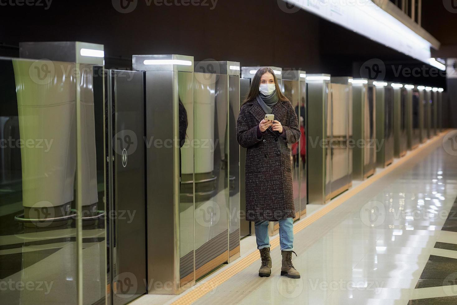 una mujer con una mascarilla médica está esperando un tren y sosteniendo un teléfono inteligente foto