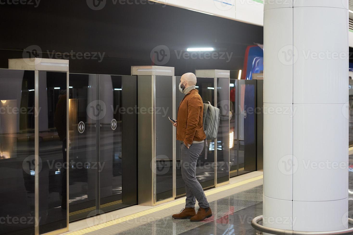 A man in a face mask is holding a smartphone while waiting for a subway train photo