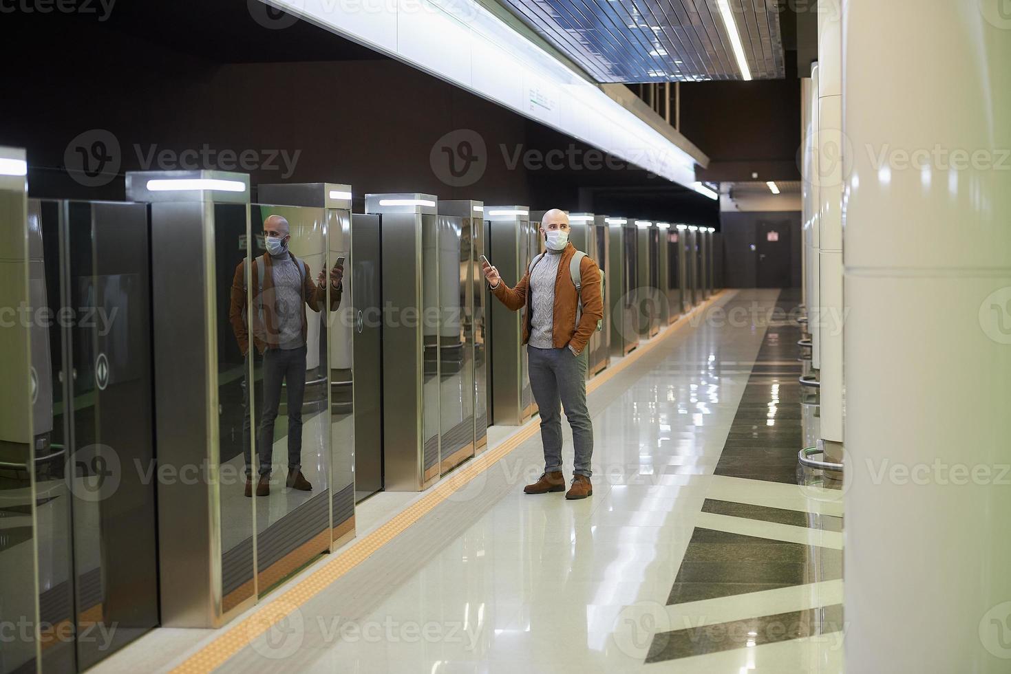 A man in a face mask is using a smartphone while waiting for a subway train photo