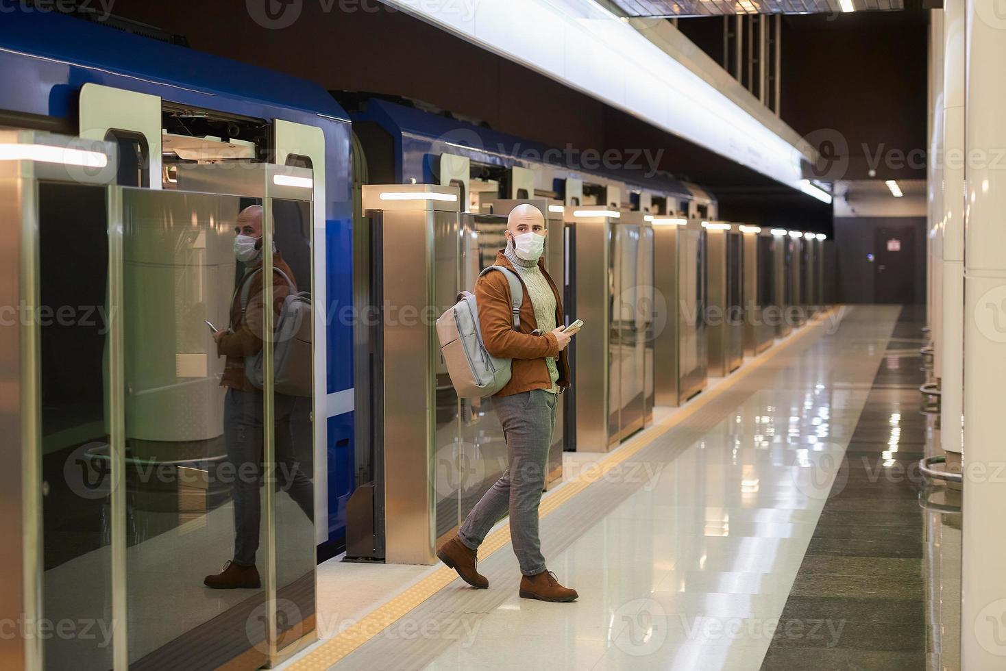 El hombre con una mascarilla médica sostiene un teléfono mientras deja un vagón de metro moderno foto