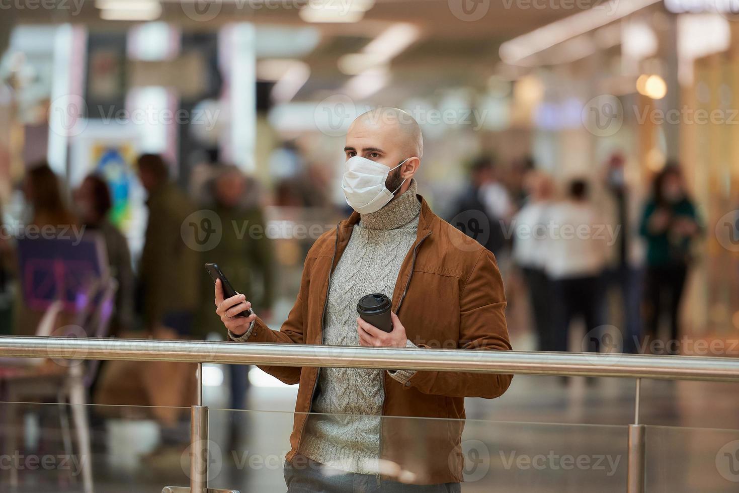 El hombre con una mascarilla está usando un teléfono y sosteniendo un café en el centro comercial. foto