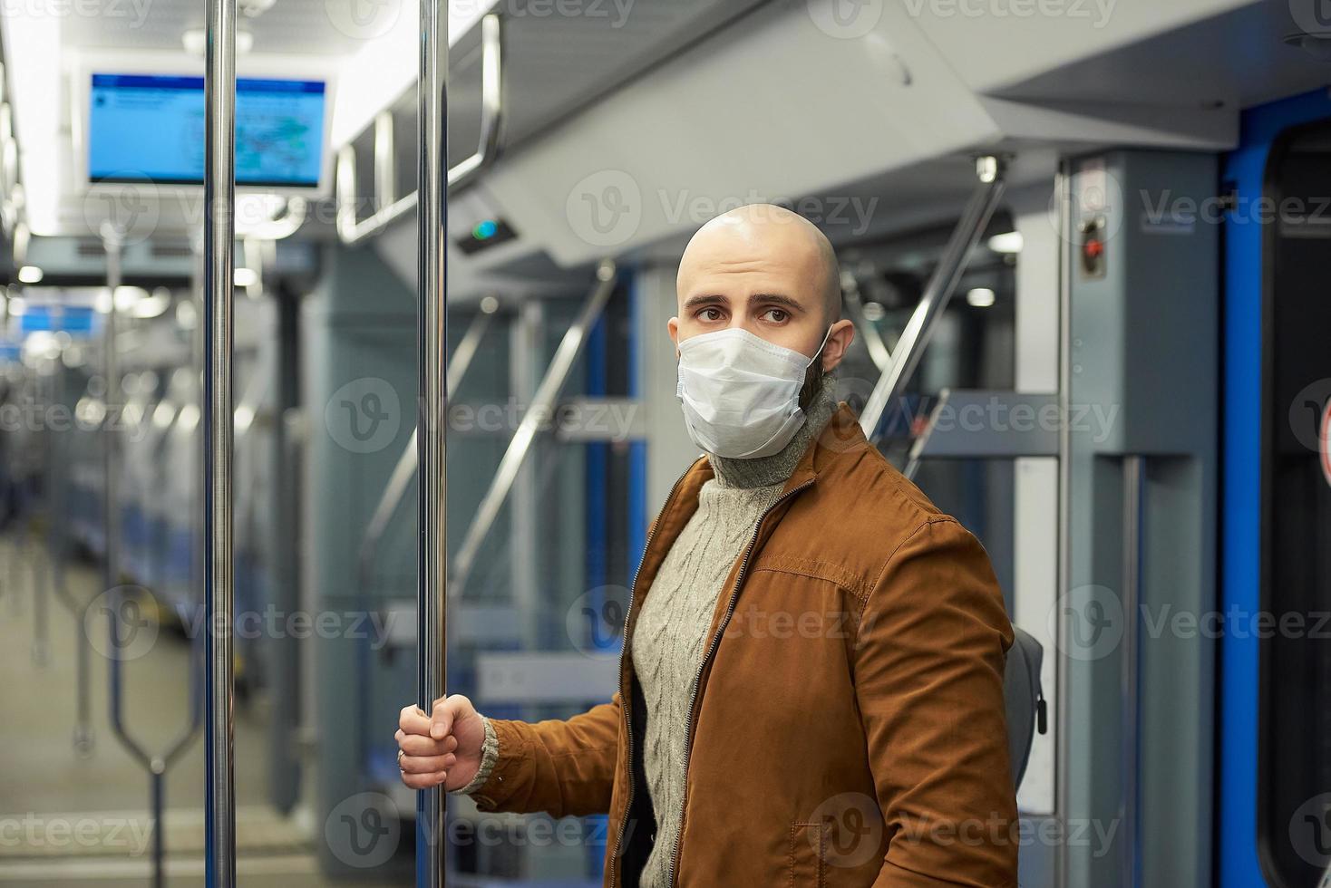 Un hombre calvo con barba en una mascarilla está sosteniendo el pasamanos en un vagón de metro foto