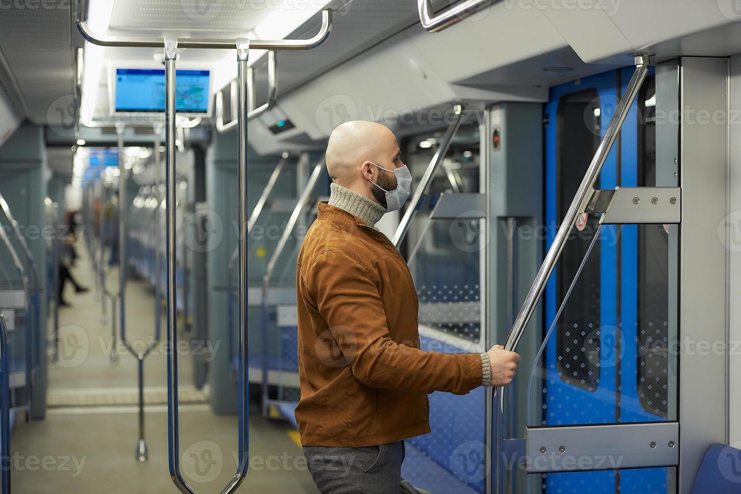 Un hombre calvo con barba en una mascarilla está sosteniendo el pasamanos en un vagón de metro foto