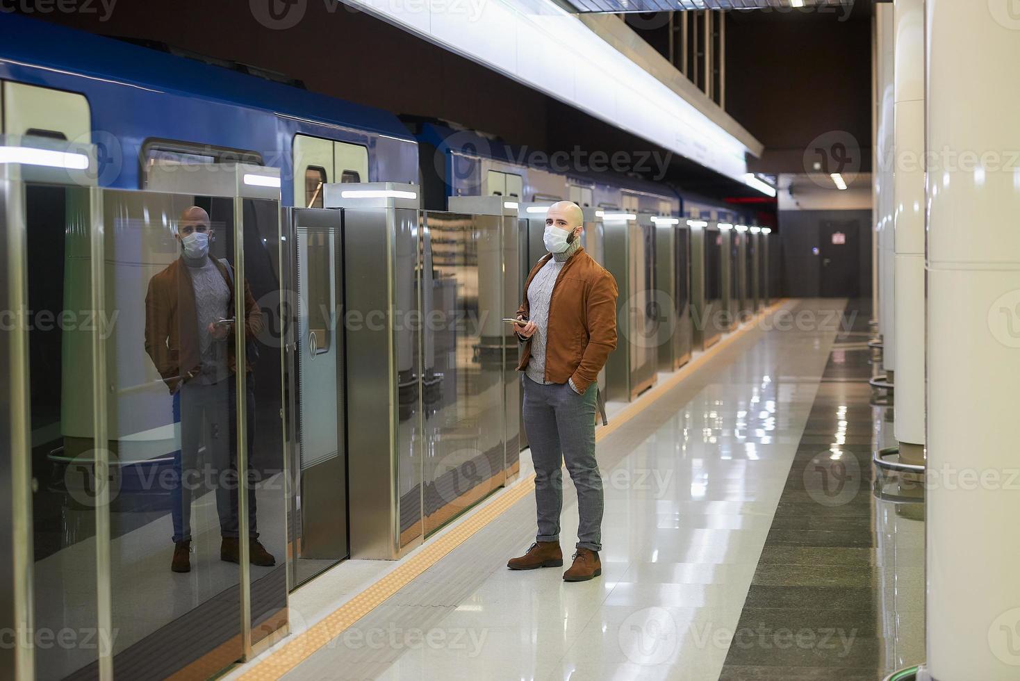 A man in a face mask is holding a smartphone while waiting for a subway train photo