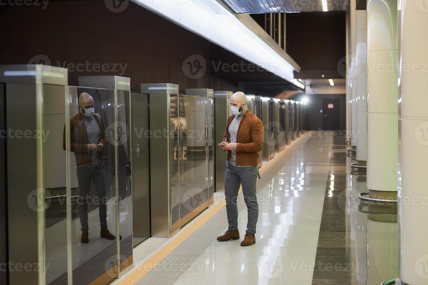 un hombre con una máscara facial está desplazando las noticias mientras espera un tren subterráneo foto