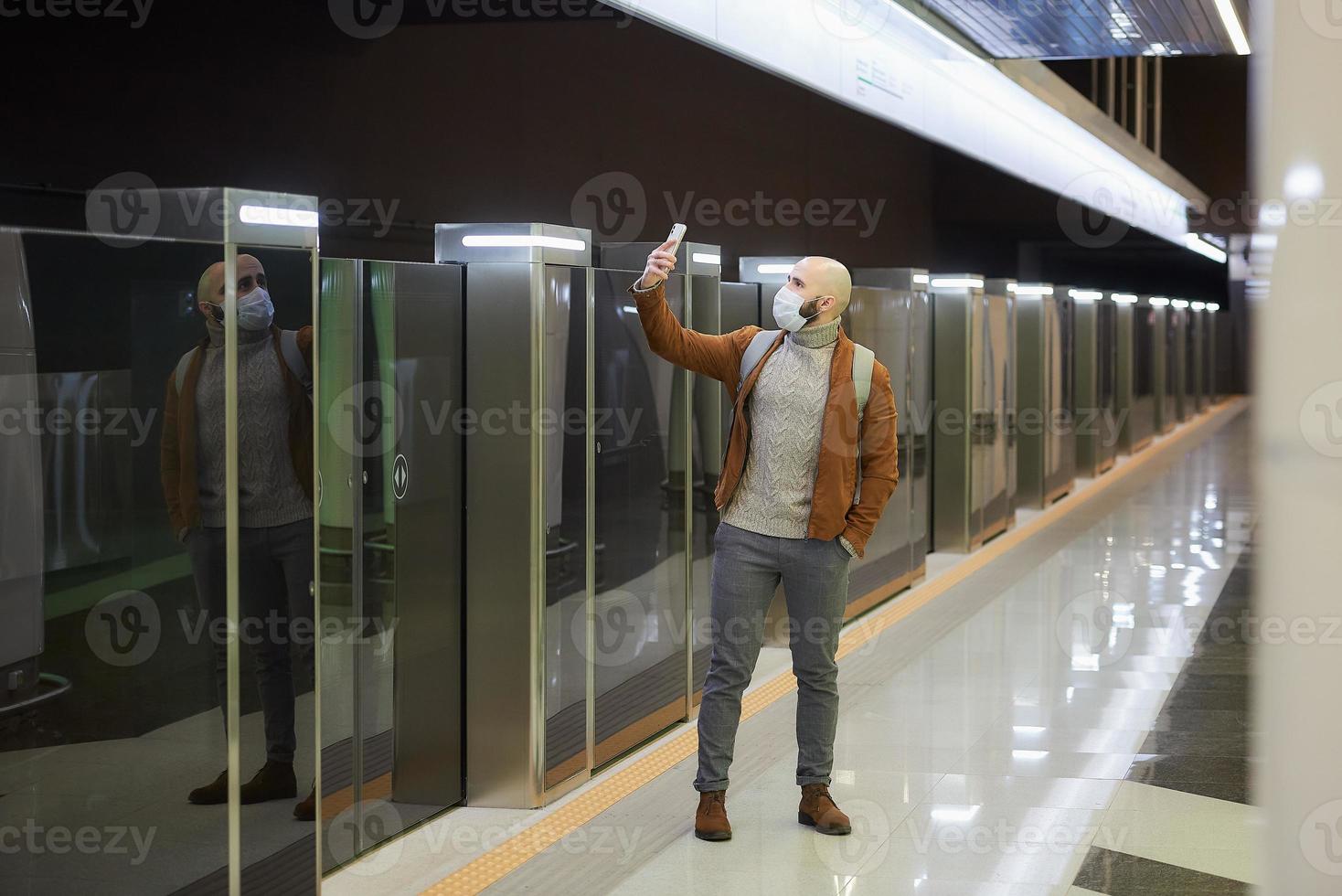 A man in a face mask is using a smartphone while waiting for a subway train photo