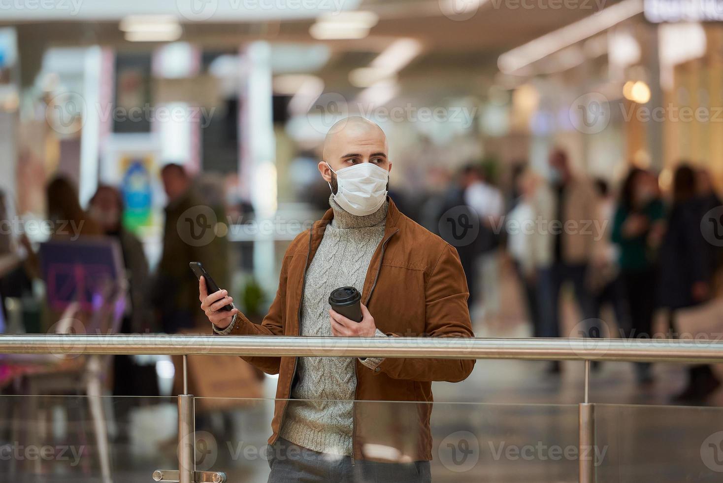 El hombre con una mascarilla está usando un teléfono y sosteniendo un café en el centro comercial. foto