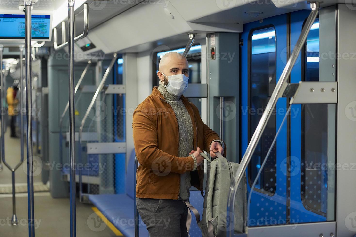 A bald man with a beard in a face mask is putting on a backpack in a subway car photo