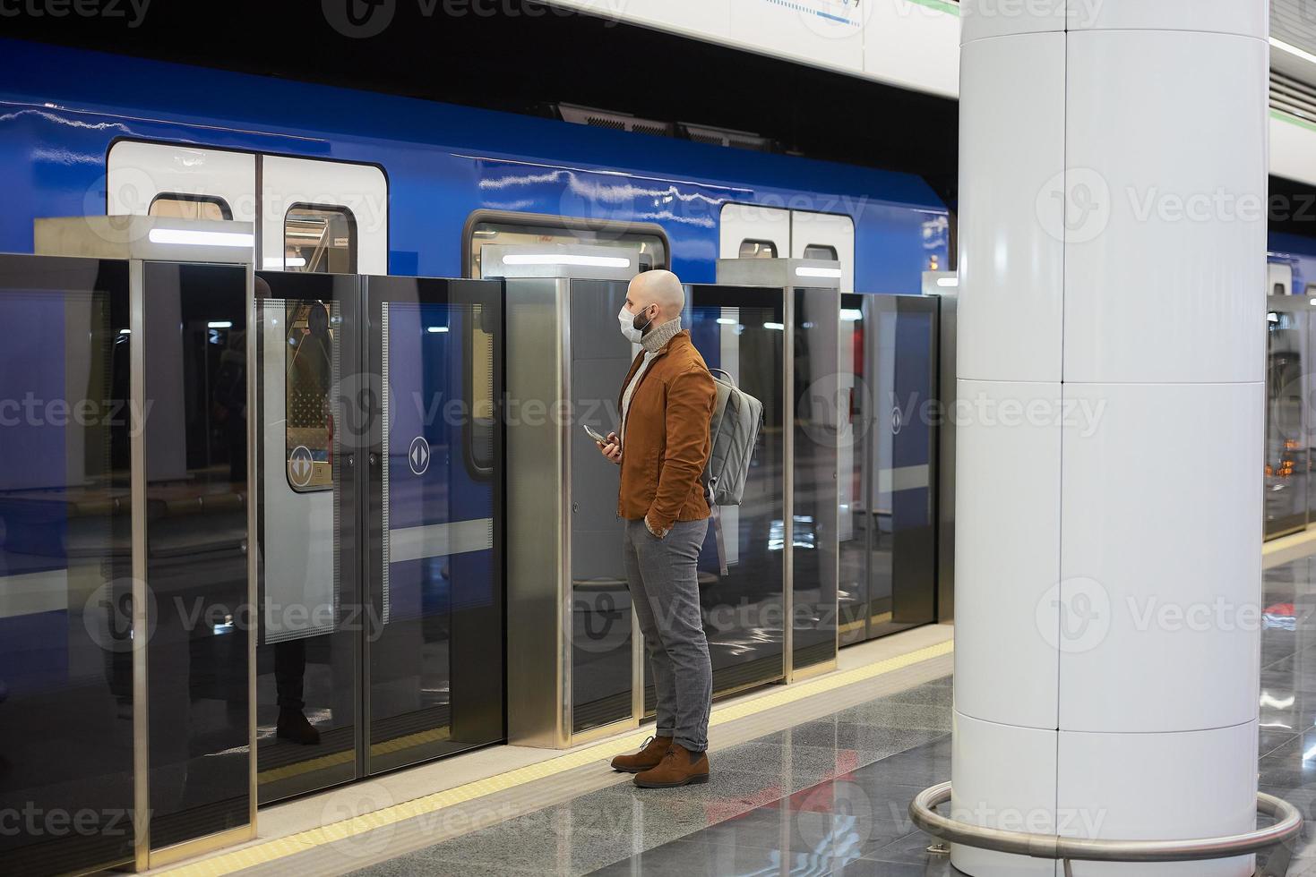 A man in a face mask is holding a smartphone while waiting for a subway train photo