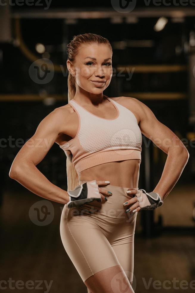 A fit woman with blonde hair is posing with her hands at her waist in a gym photo