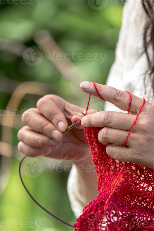 Hands of a woman who crochets with red wool knits in front of blurred background photo