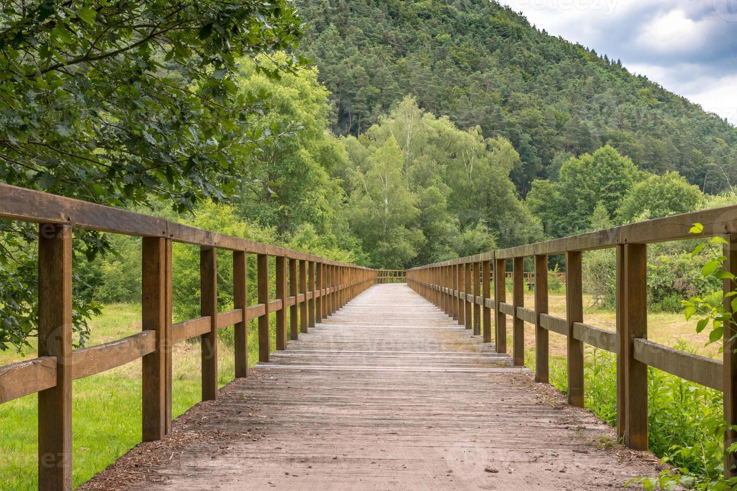 Puente de madera sobre un campo con prados y colinas boscosas foto