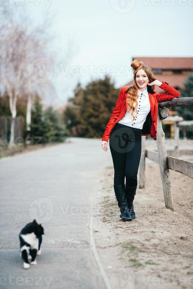 Red-haired jockey girl in a red cardigan and black high boots with a farm cat photo