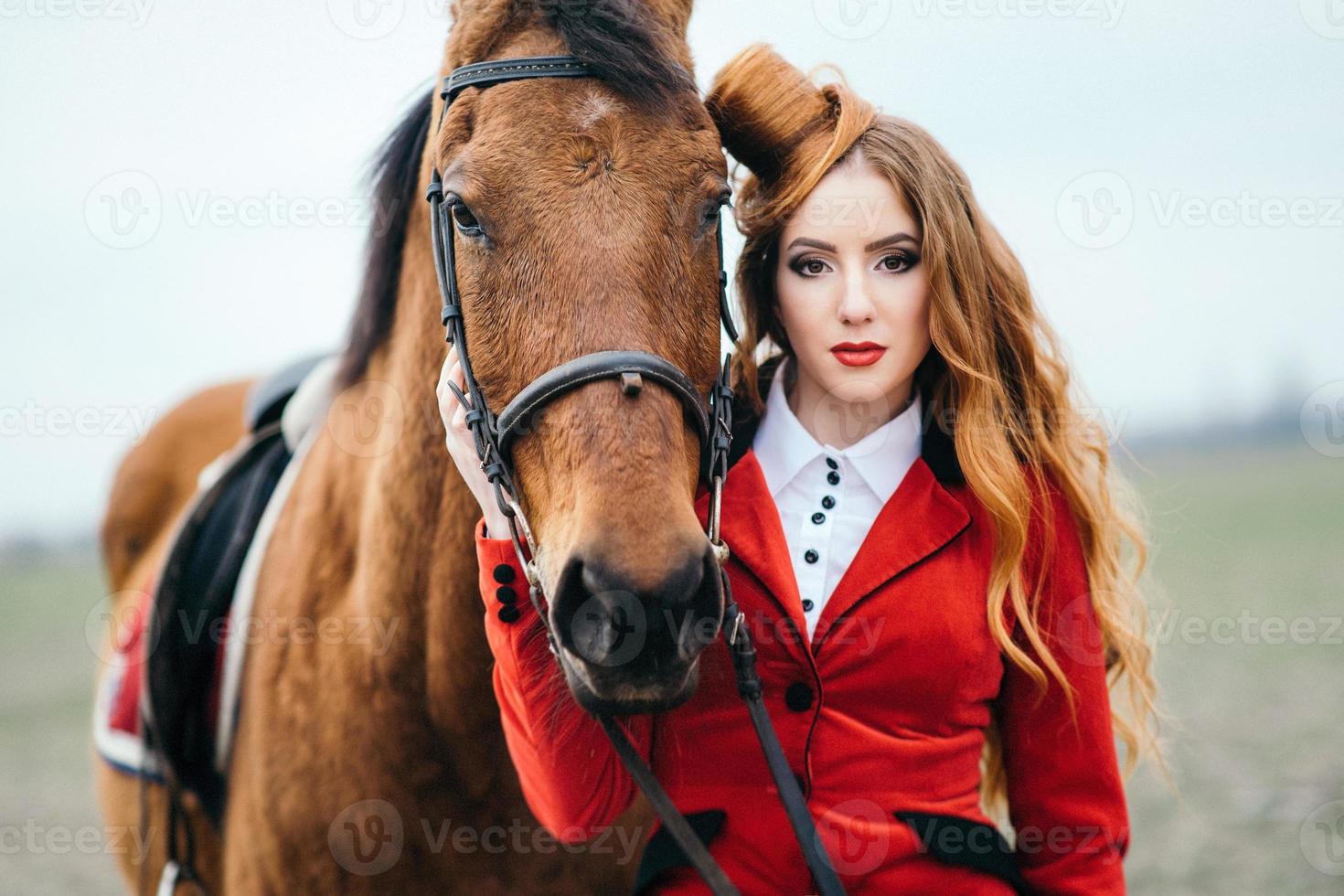 Red-haired jockey girl in a red cardigan and black high boots with a horse photo