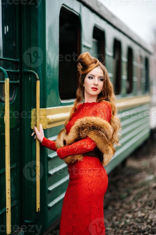 Chica joven con cabello rojo con un vestido rojo brillante cerca de un viejo automóvil de pasajeros foto