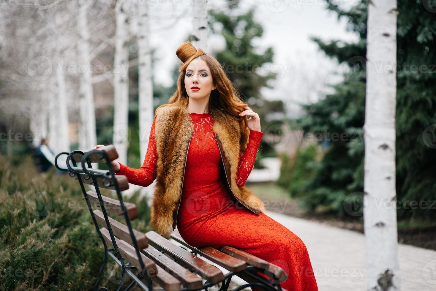 Chica joven con el pelo rojo con un vestido rojo brillante en un banco en un parque vacío foto