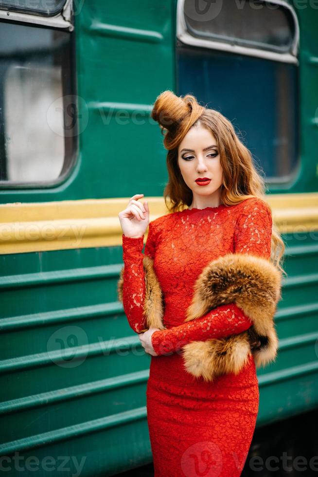 Chica joven con cabello rojo con un vestido rojo brillante cerca de un viejo automóvil de pasajeros foto