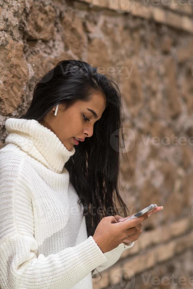 Hermosa joven morena en ropa de invierno con su teléfono inteligente escuchando música con auriculares foto