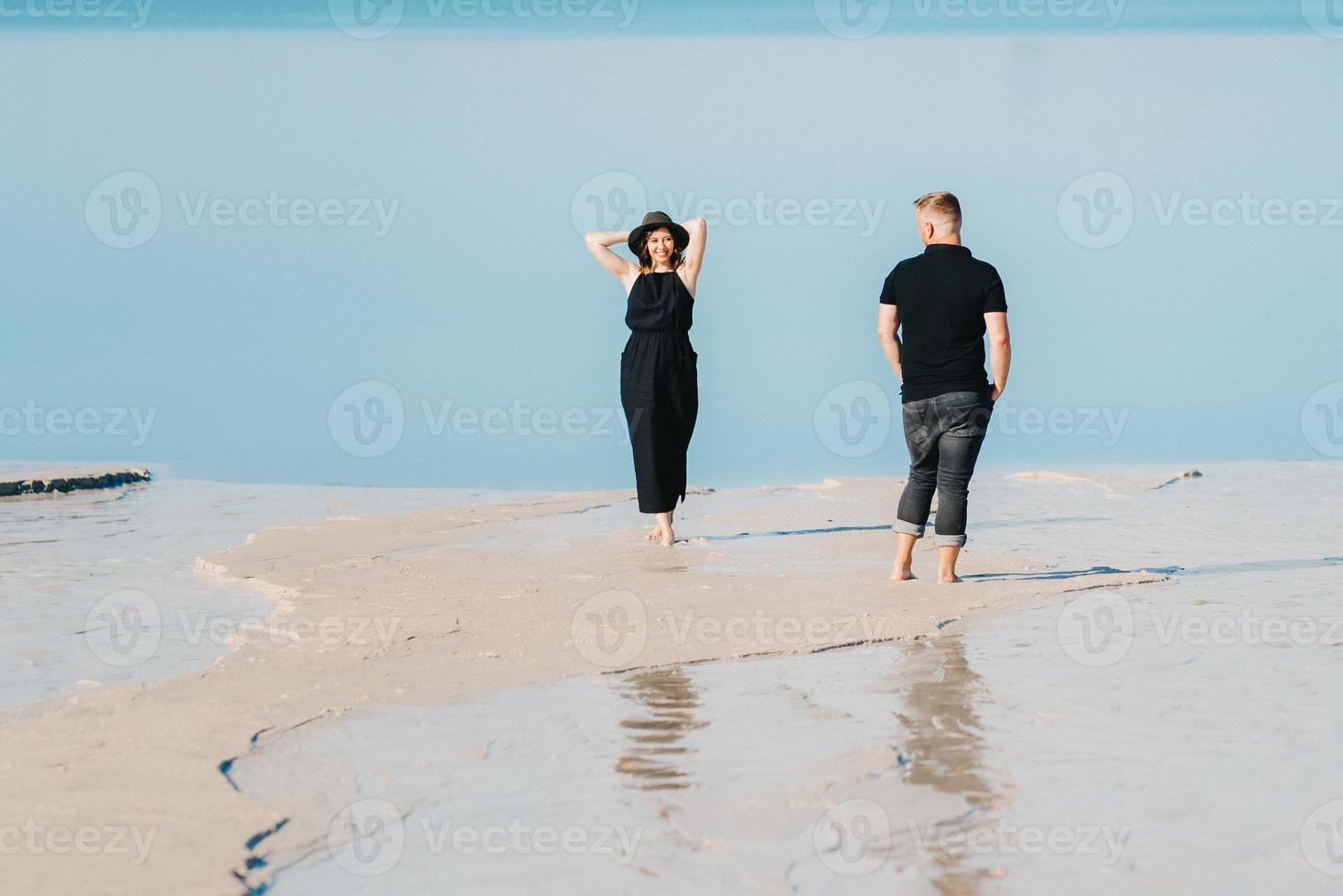 young couple a guy with a girl in black clothes are walking on the white sand photo