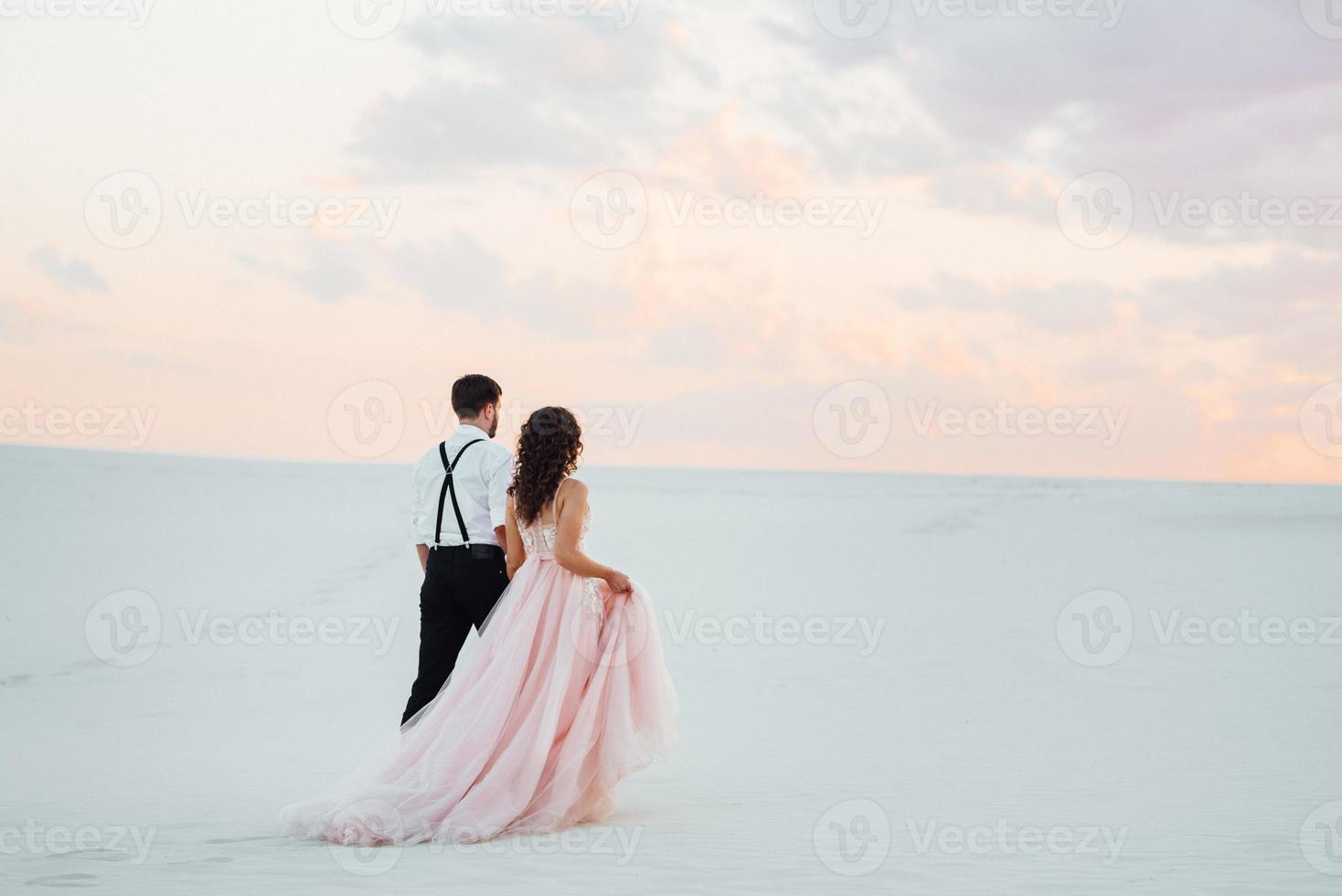 young couple a guy in black breeches and a girl in a pink dress are walking along the white sand photo