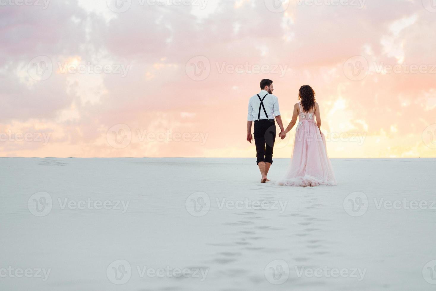 young couple a guy in black breeches and a girl in a pink dress are walking along the white sand photo