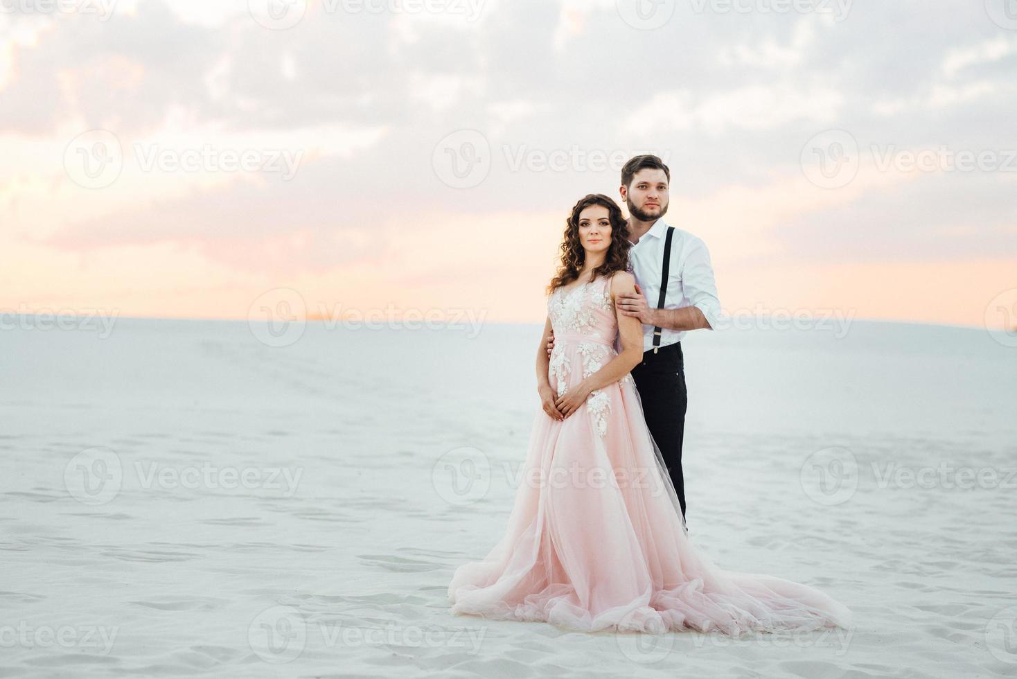 young couple a guy in black breeches and a girl in a pink dress are walking along the white sand photo