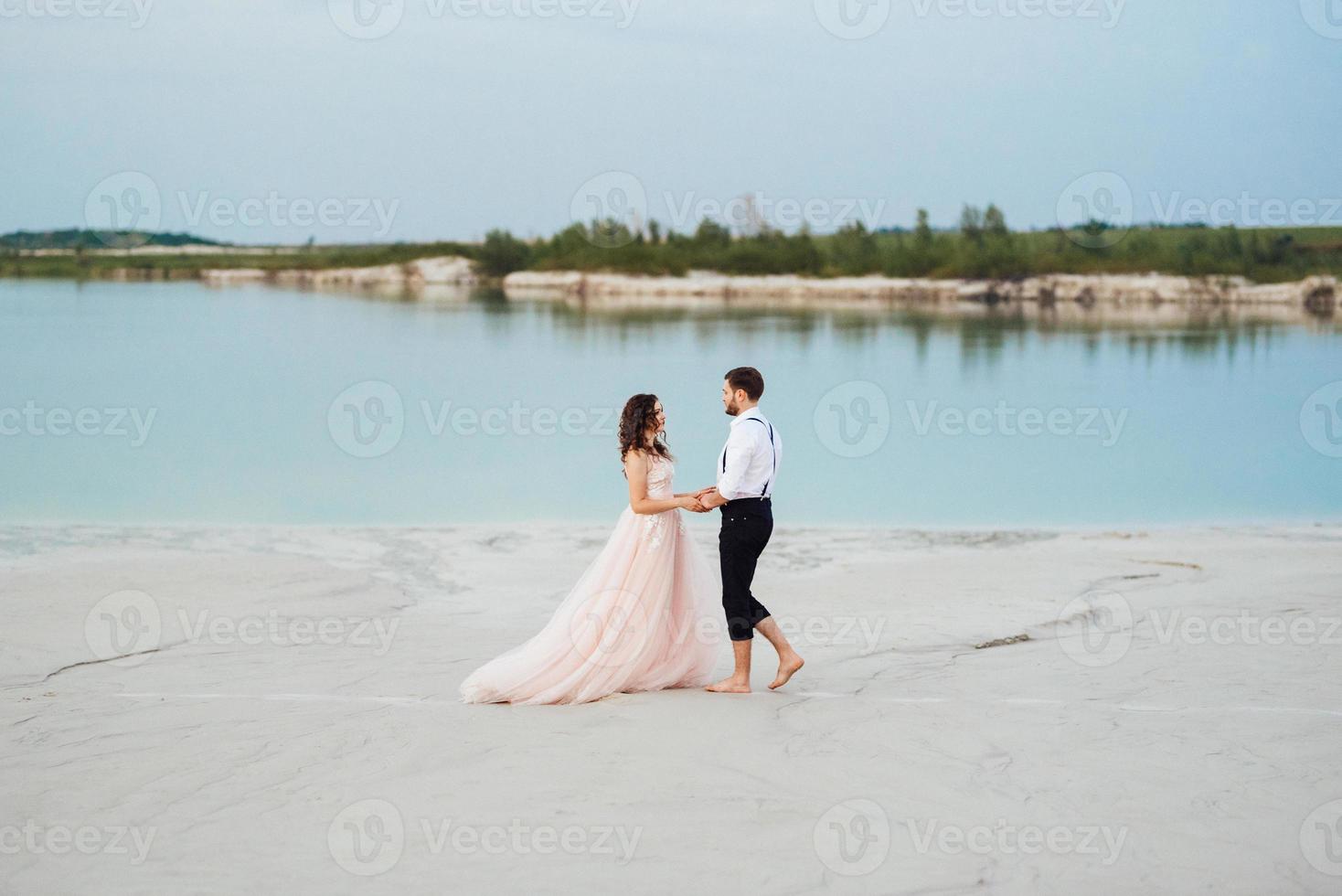 Pareja joven un chico con pantalones negros y una chica con un vestido rosa están caminando por la arena blanca foto