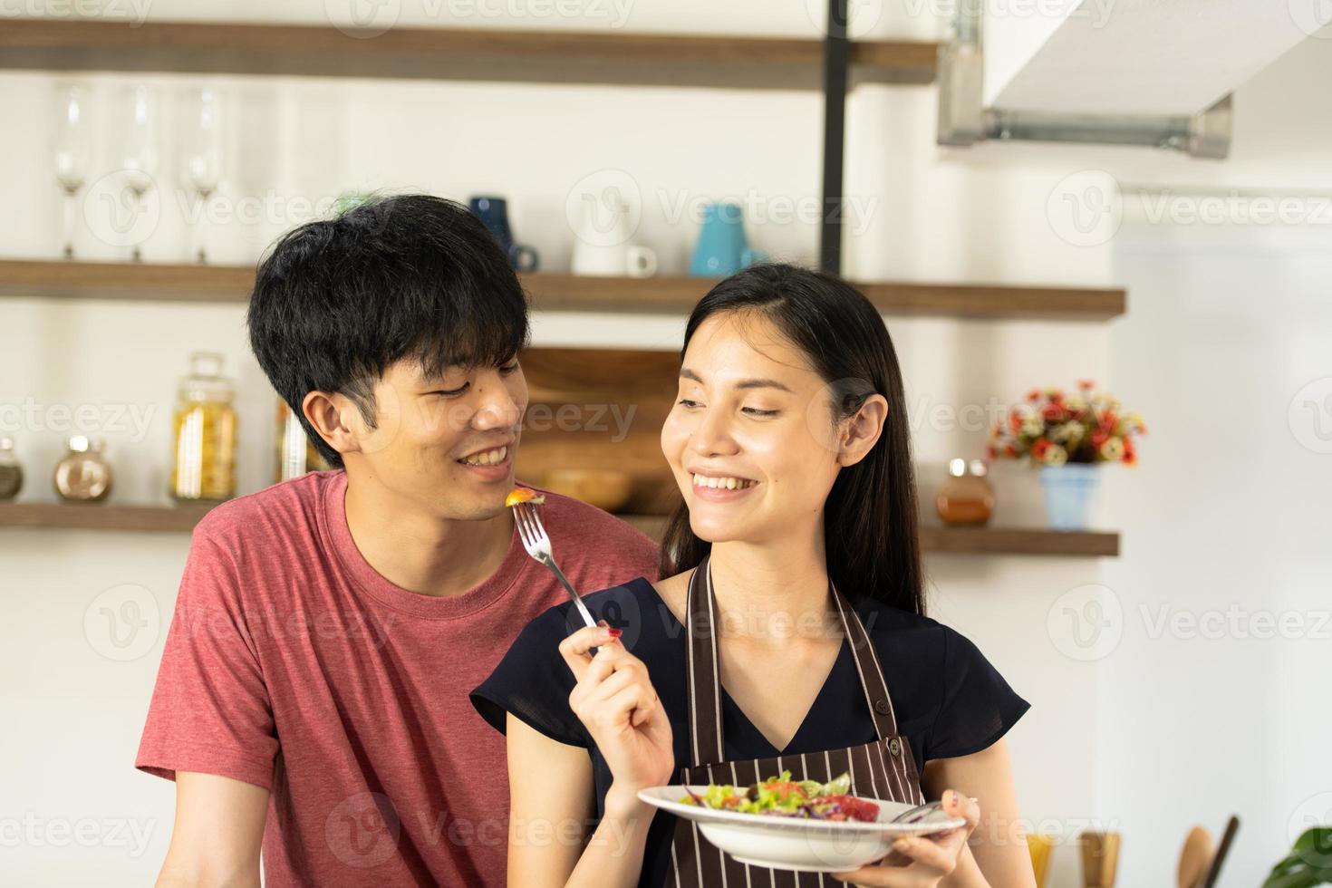 A young Asian couple are eating together and smiling happily while cooking their salad in the kitchen. photo