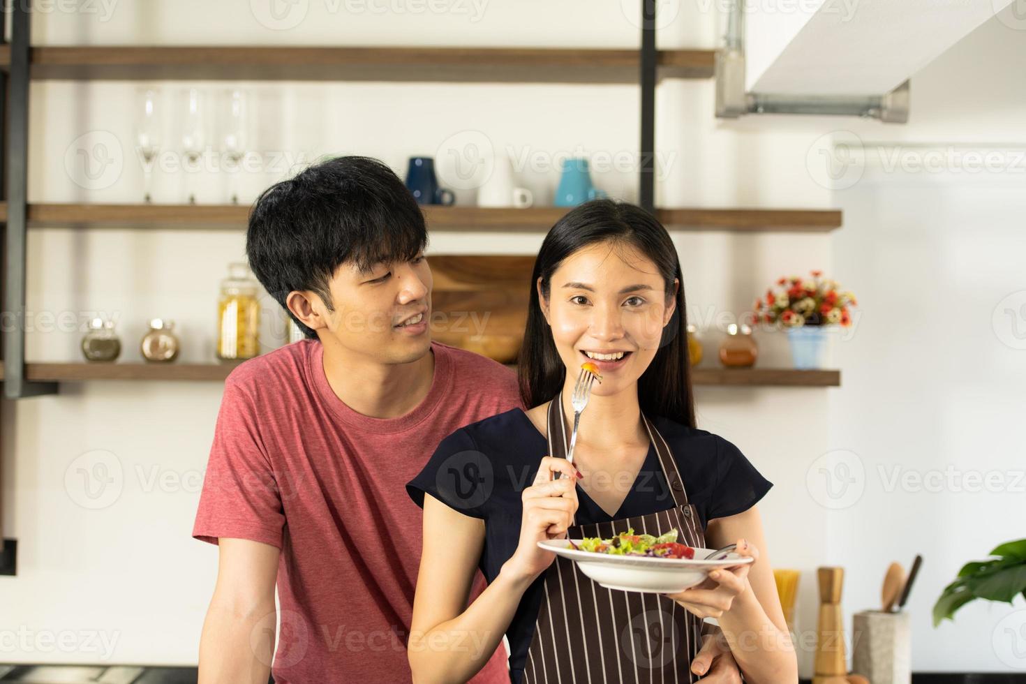 A young Asian couple are eating together and smiling happily while cooking their salad in the kitchen. photo