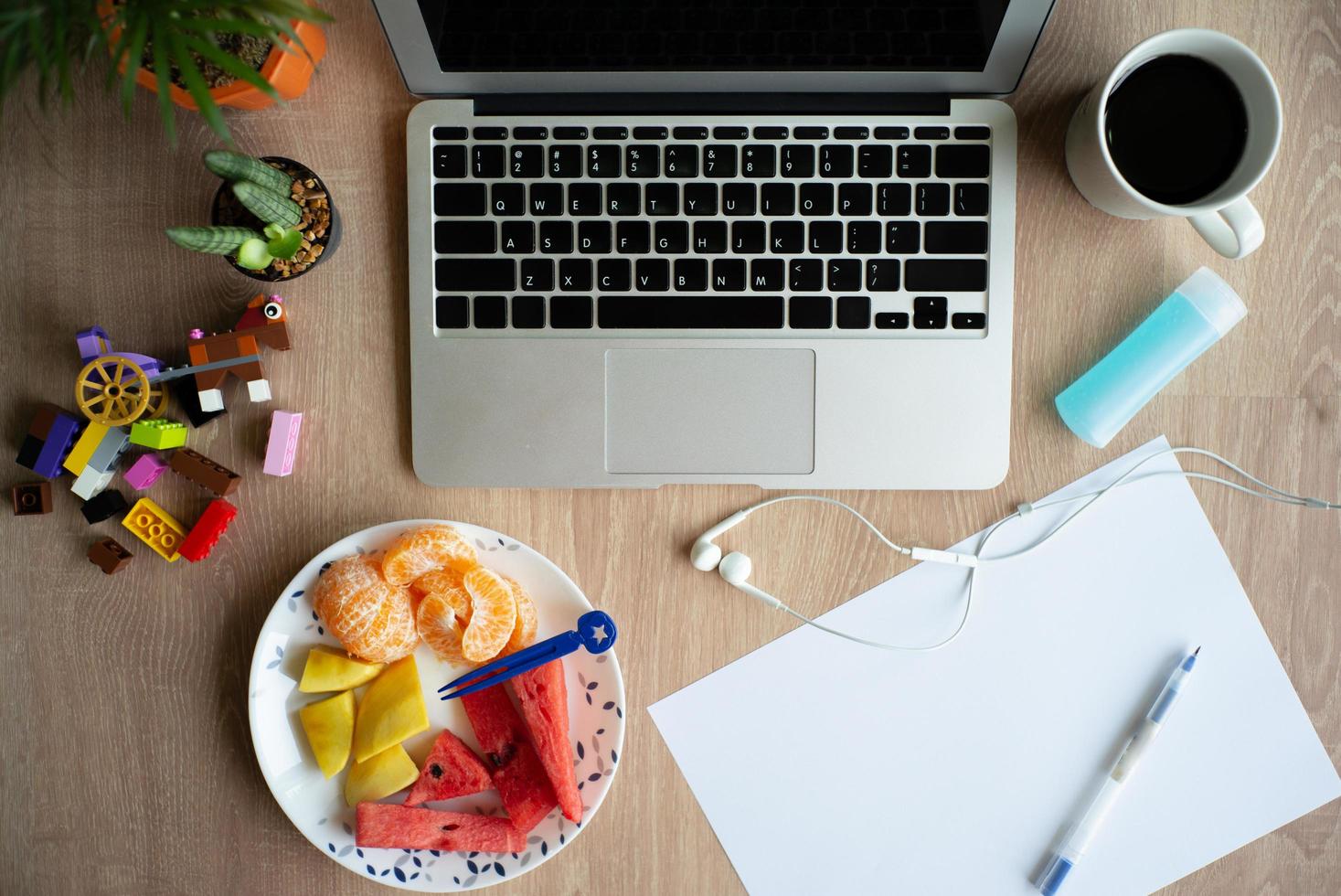 Vista superior de una computadora portátil en el escritorio de madera con papelería, café y frutas mixtas en un plato blanco. trabajar desde casa. cuarentena en casa foto