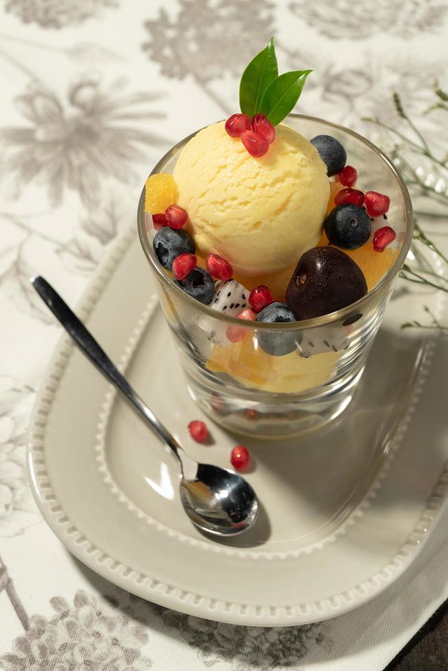 Top view of vanilla ice cream in a glass bowl topped with mixed fruits and decorated by a small leaf photo