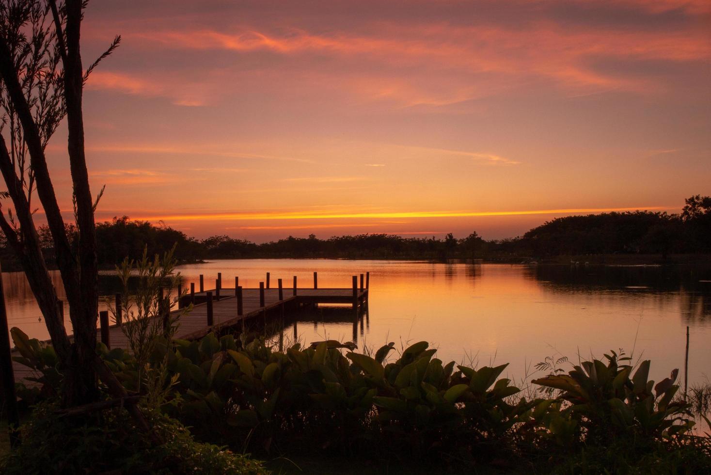 Pasarela de madera en el lago con un paisaje natural de puesta de sol y silueta de bosque en segundo plano. foto