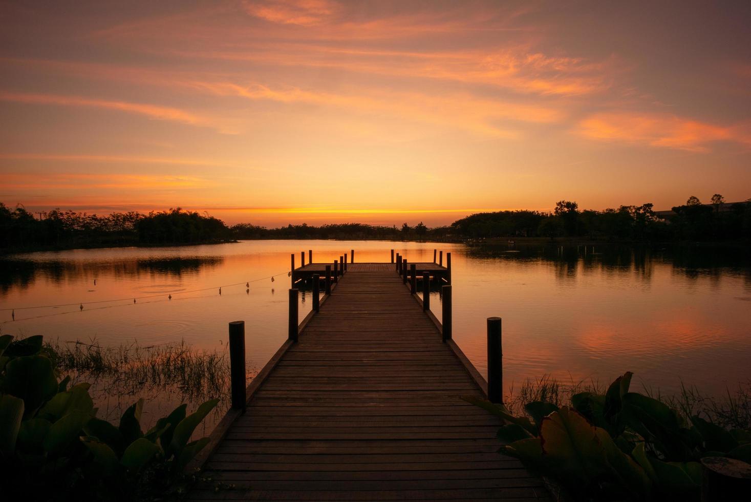 Pasarela de madera en el lago con un paisaje natural de puesta de sol y silueta de bosque en segundo plano. foto