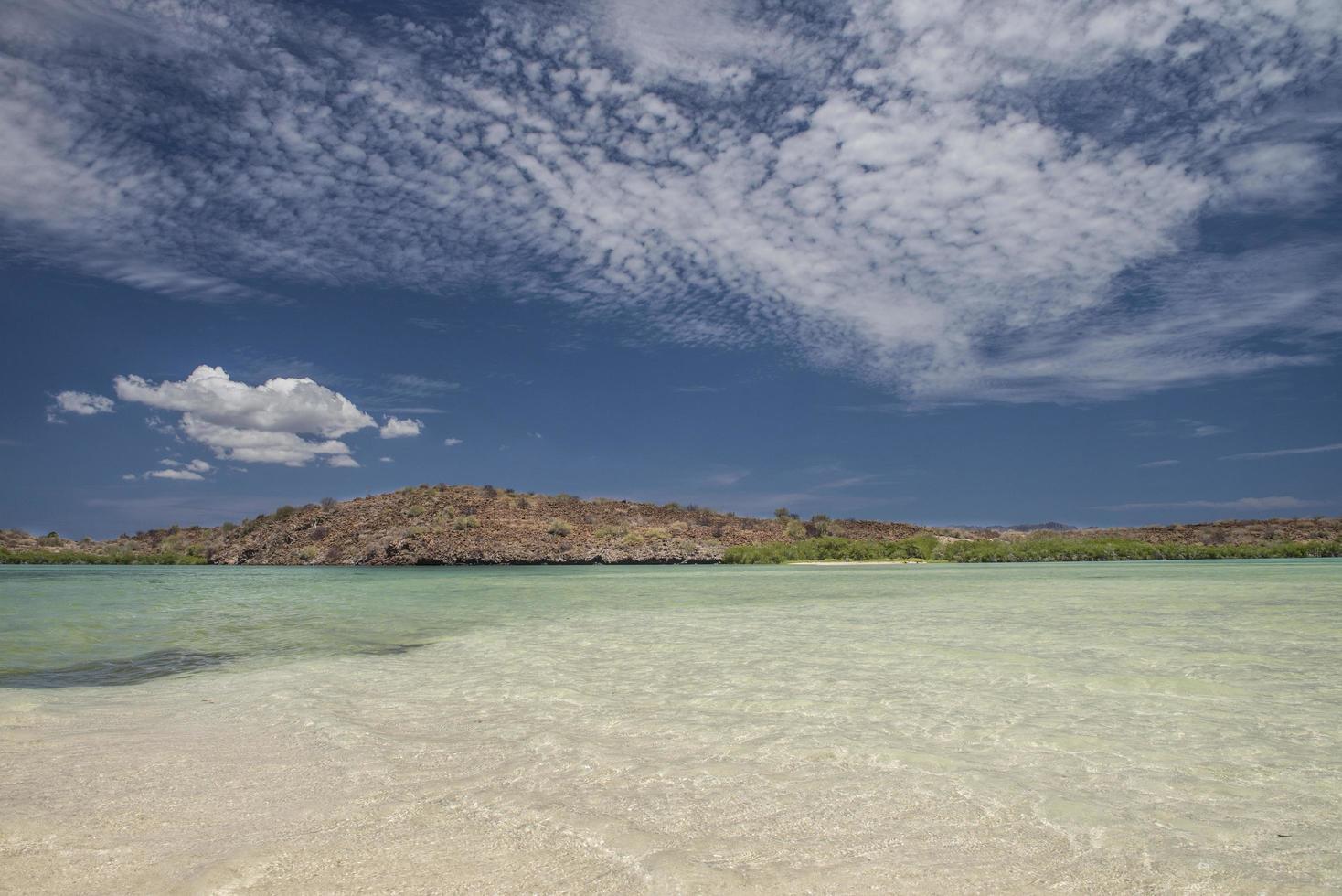 Agua de mar clara con montañas en el fondo y cielo azul con nubes en la península de Baja, Baja California foto