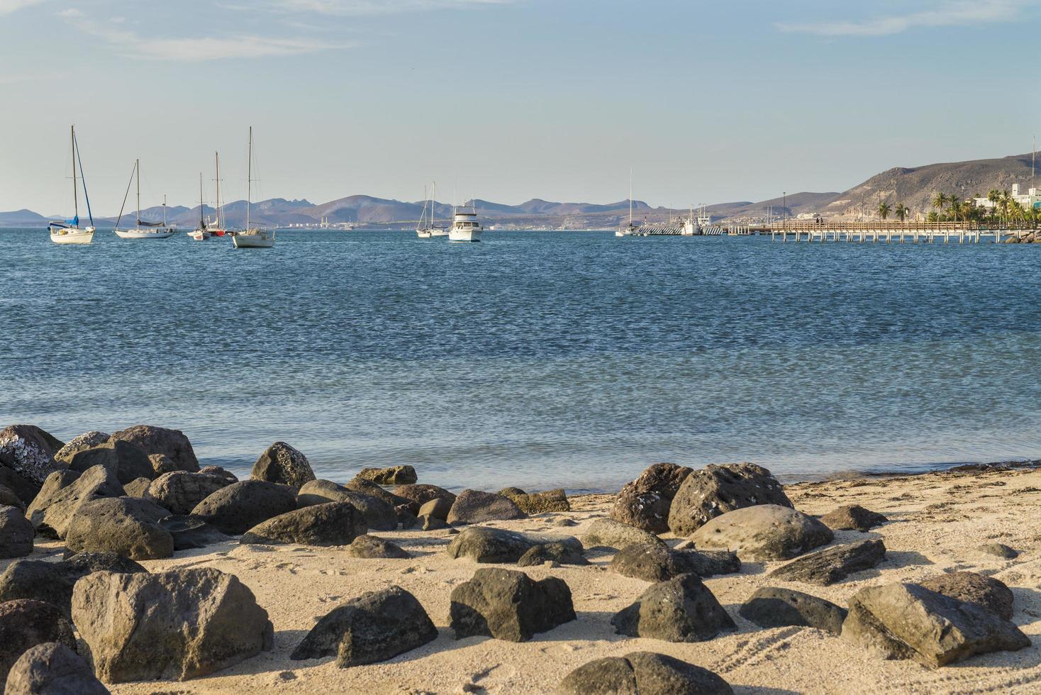 malecón de la paz por el mar de cortes con rocas en la playa y una vista de la ciudad con barcos en el mar foto