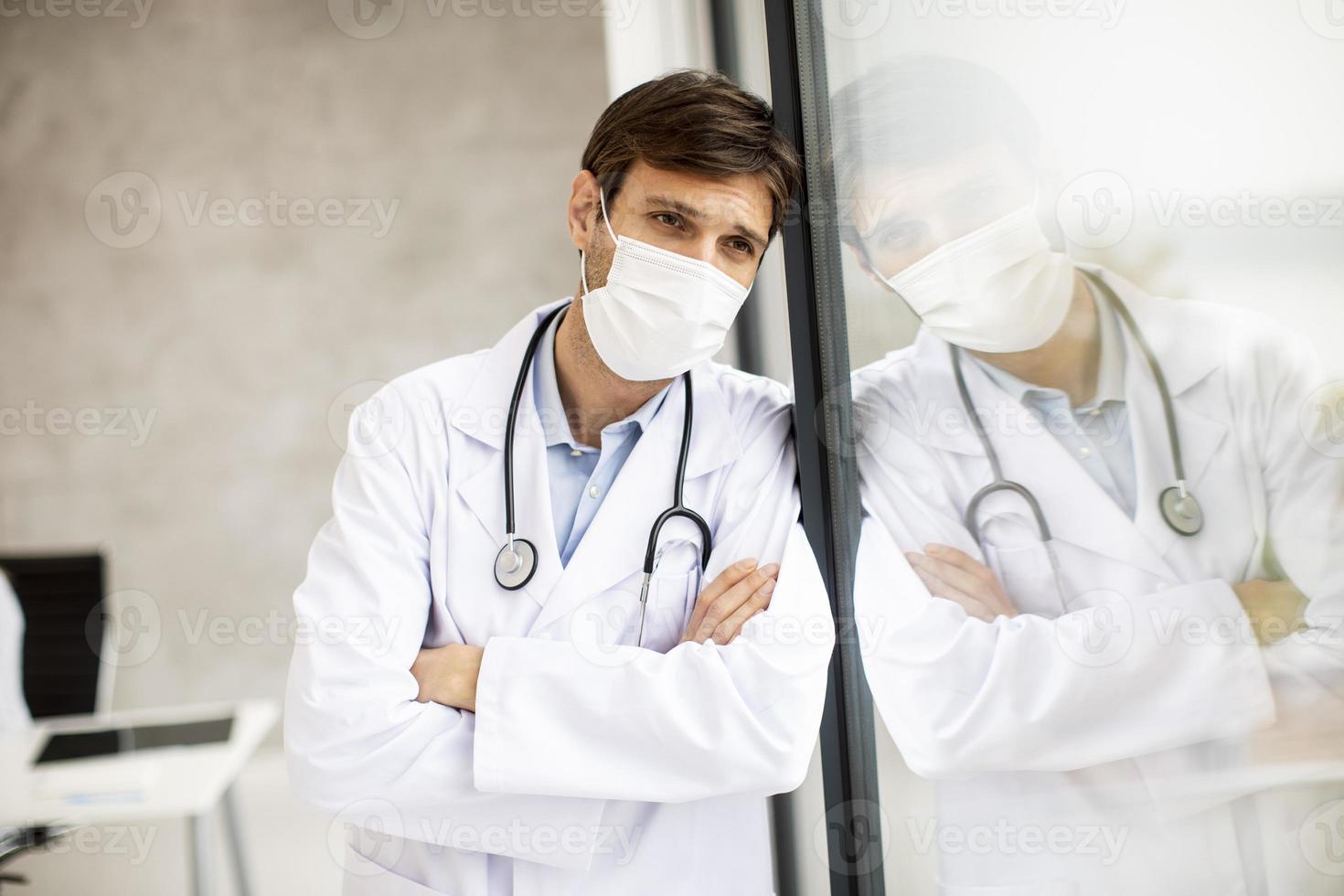 Doctor leaning against a window while wearing a mask photo