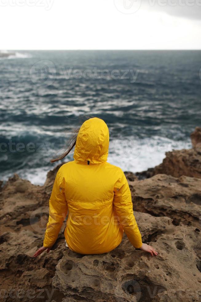 Mujer joven vestida con impermeable amarillo sentado en el acantilado mirando grandes olas foto