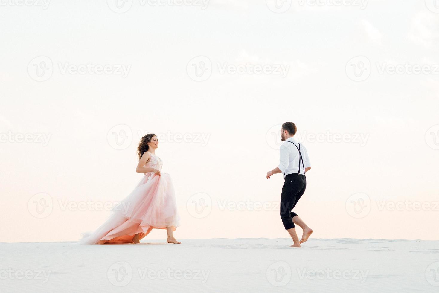 Pareja joven un chico con pantalones negros y una chica con un vestido rosa están caminando por la arena blanca foto