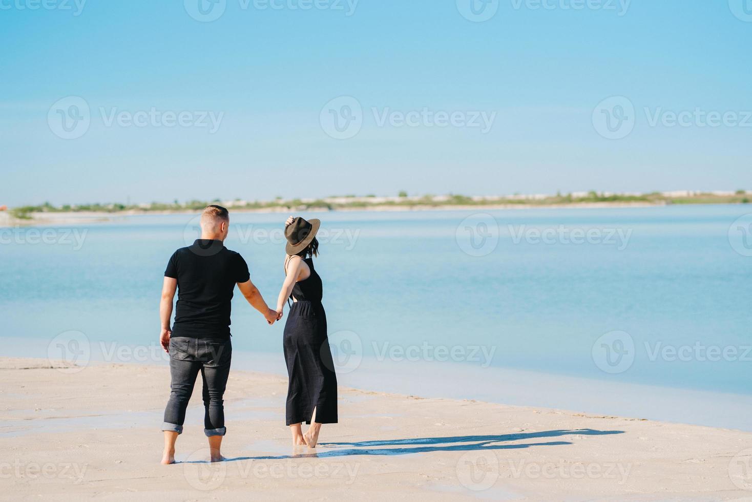 Young couple a guy with a girl in black clothes are walking on the white sand photo