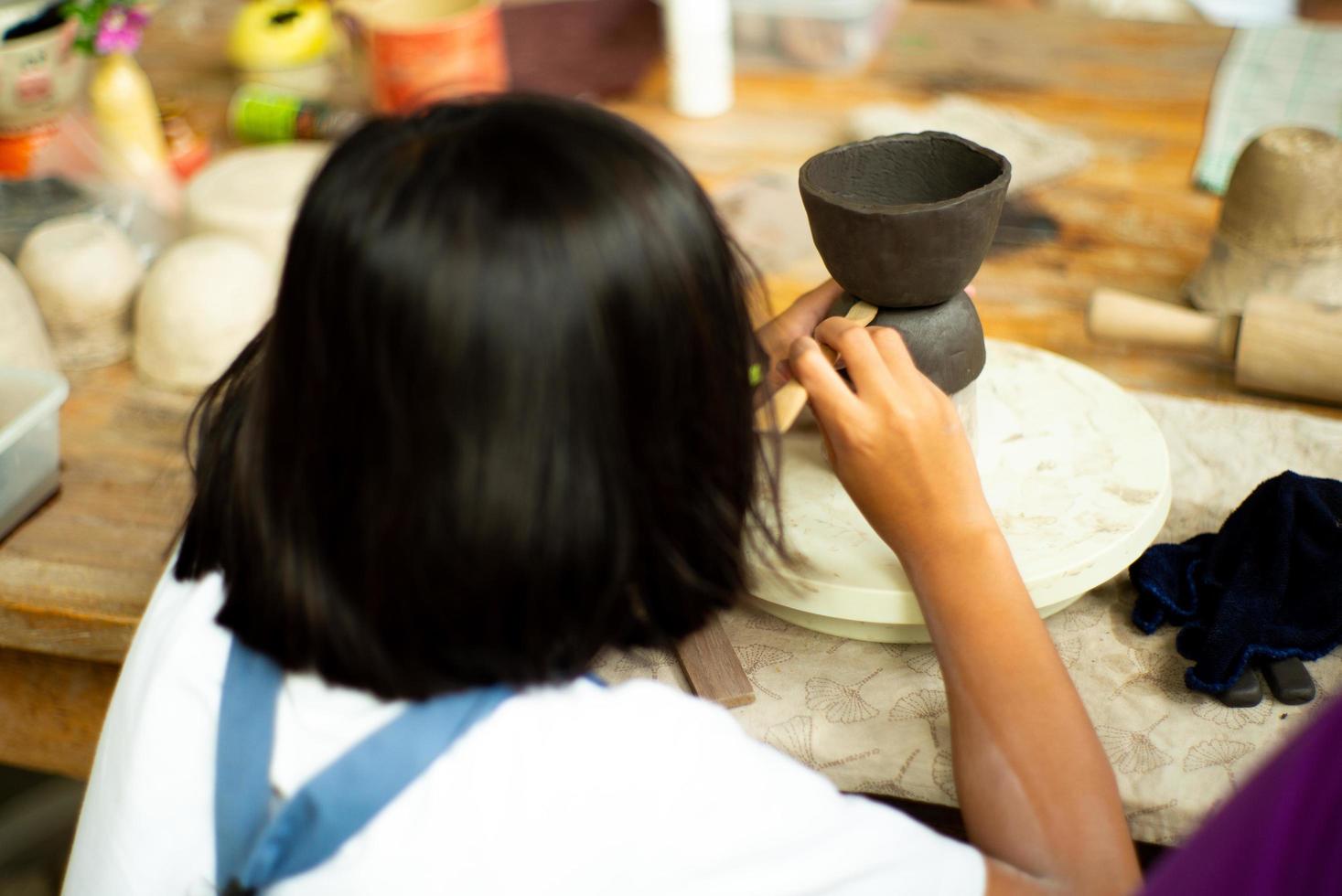 Primer plano de las manos de la niña moldeando la arcilla en la bandeja de la rueda con un palo de madera con la espalda borrosa retrato de niña en primer plano foto