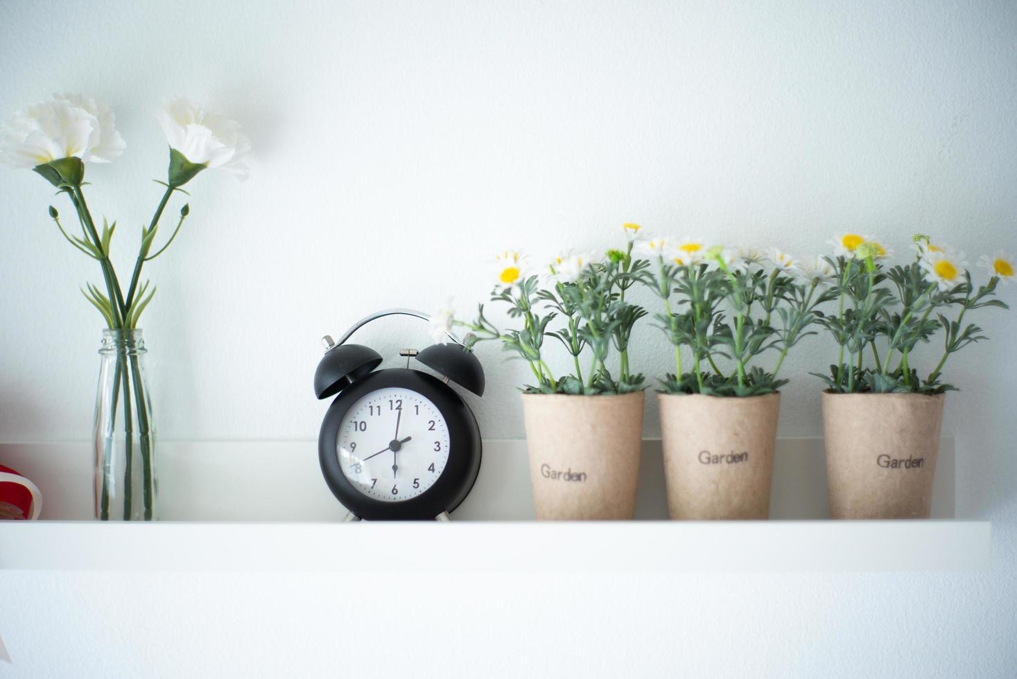 Vintage alarm clock on the white wooden shelf decorated by the blossom flowers in the pottery with blurred background of the vase. photo